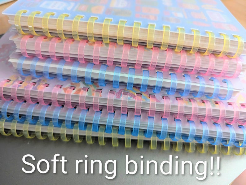May include: A stack of notebooks with soft ring binding. The notebooks have clear plastic covers and colourful spiral bindings in yellow, pink, and blue. The text "Soft ring binding!!" is visible at the bottom of the image.