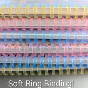May include: Close-up of a stack of notebooks with soft ring binding. The spiral bindings are in yellow, pink, and blue. The text "Soft Ring Binding!" is at the bottom of the image.