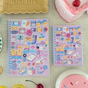 May include: Two spiral-bound notebooks with a light purple cover featuring retro-themed stickers. The stickers include arcade games, roller skates, and computer graphics. The text "BE KIND REWIND TOTALLY" is visible. A decorative biscuit and a heart-shaped cake are also in the image.