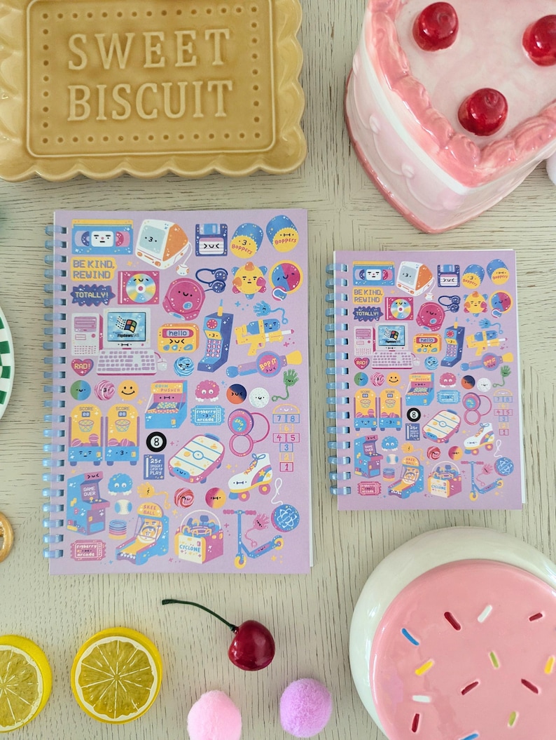 May include: Two spiral-bound notebooks with a light purple cover featuring retro-themed stickers. The stickers include arcade games, roller skates, and computer graphics. The text "BE KIND REWIND TOTALLY" is visible. A decorative biscuit and a heart-shaped cake are also in the image.