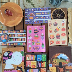 K&ouml;nnte beinhalten: Eine Sammlung von Aufklebern und Dekorationsartikeln mit Herbst- und Halloween-Motiven. Enth&auml;lt Aufkleber mit Geister-, Katzen- und K&uuml;rbisdesigns sowie einen Keramikteller und eine getrocknete Orangenscheibe. Die Aufkleber enthalten Text wie "Treats Only".