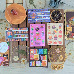 K&ouml;nnte beinhalten: Eine Sammlung dekorativer Aufkleber, Untersetzer und eines Keramikst&uuml;cks. Die Aufkleber zeigen verschiedene Designs, darunter B&uuml;cher, Halloween-Leckereien und Kaffee. Die Untersetzer sind aus Harz gefertigt und zeigen getrocknete Blumen. Das Keramikst&uuml;ck ist rund und hat eine rote Beere.