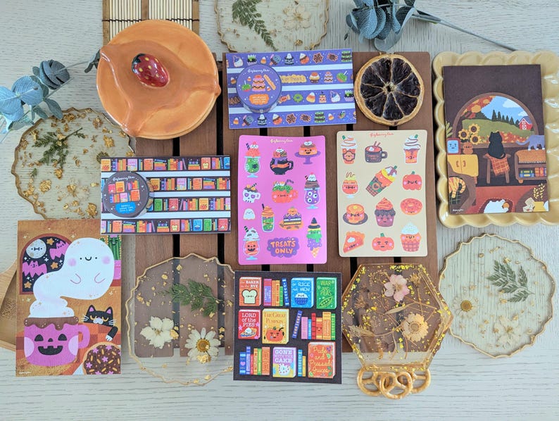 K&ouml;nnte beinhalten: Eine Sammlung dekorativer Aufkleber, Untersetzer und eines Keramikst&uuml;cks. Die Aufkleber zeigen verschiedene Designs, darunter B&uuml;cher, Halloween-Leckereien und Kaffee. Die Untersetzer sind aus Harz gefertigt und zeigen getrocknete Blumen. Das Keramikst&uuml;ck ist rund und hat eine rote Beere.