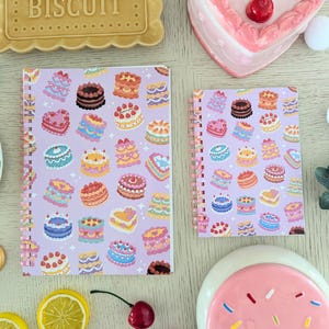 May include: Two spiral-bound notebooks with a lavender cover featuring colorful illustrations of cakes. A ceramic biscuit with the text "SWEET BISCUIT" is at the top. Other items include a heart-shaped dish with cherries, lemon slices, and decorative elements.