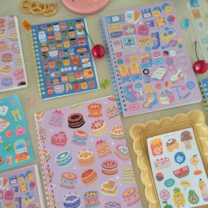 May include: A collection of spiral-bound notebooks and stickers with colorful illustrations. The notebooks feature designs of cakes, food items, and retro electronics. A pink plate with sprinkles and a yellow picture frame are also visible.