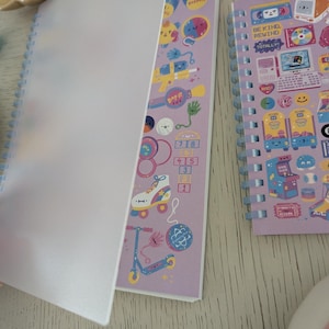 May include: Two spiral-bound notebooks with colourful illustrations. One notebook is open, revealing pages with playful graphics of toys and the text "Bop It." The other notebook features retro gaming and the text "Be Kind, Rewind."