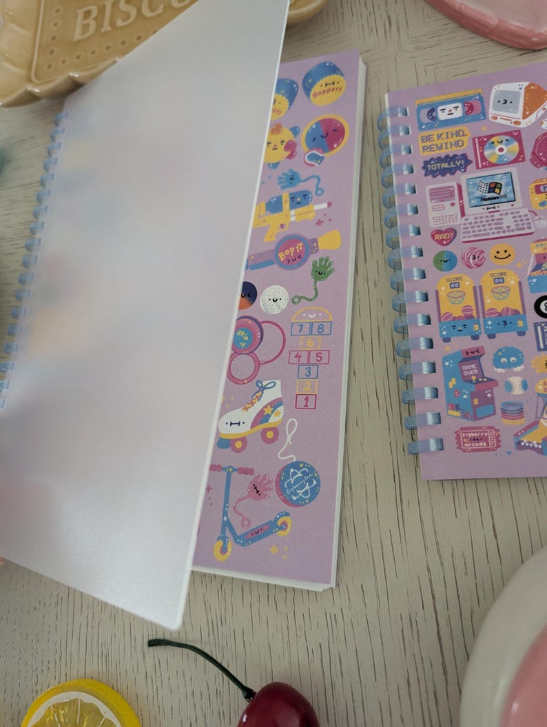 May include: Two spiral-bound notebooks with colourful illustrations. One notebook is open, revealing pages with playful graphics of toys and the text "Bop It." The other notebook features retro gaming and the text "Be Kind, Rewind."