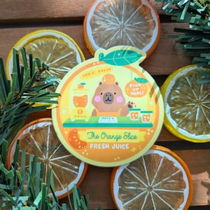 Peut inclure: Un autocollant circulaire avec un personnage d'ours et le texte "The Orange Slice FRESH JUICE" est entouré de tranches d'orange et de citron séchées. Des brins verts sont placés autour de l'autocollant et des tranches de fruits.