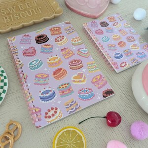 May include: Two spiral-bound notebooks with a lavender cover featuring colorful cake illustrations. The image includes a ceramic biscuit-shaped tray, a slice of cake on a plate, and decorative elements like a cherry and lemon slice.