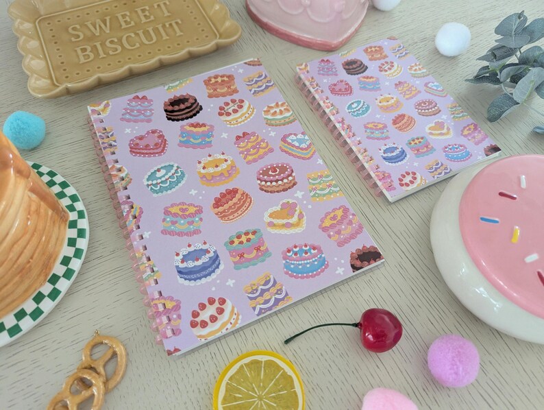 May include: Two spiral-bound notebooks with a lavender cover featuring colorful cake illustrations. The image includes a ceramic biscuit-shaped tray, a slice of cake on a plate, and decorative elements like a cherry and lemon slice.