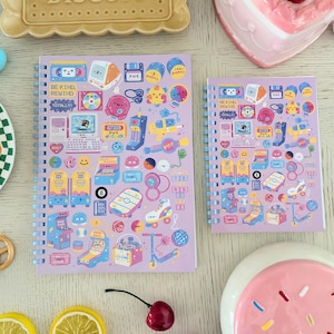 May include: Two spiral-bound notebooks with a light purple cover featuring colourful retro-themed illustrations. The notebooks have a variety of images, including computers, arcade games, and cassette tapes. The text "BE KIND REWIND" is visible on the larger notebook.