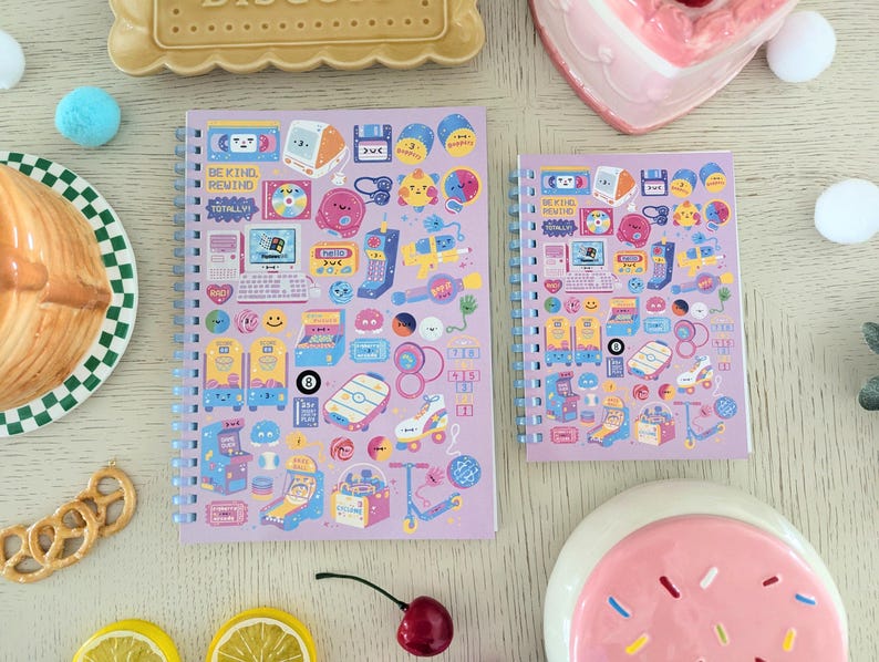 May include: Two spiral-bound notebooks with a light purple cover featuring colourful retro-themed illustrations. The notebooks have a variety of images, including computers, arcade games, and cassette tapes. The text "BE KIND REWIND" is visible on the larger notebook.