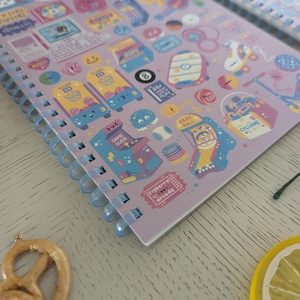 May include: A lilac spiral-bound notebook with a playful design of arcade game illustrations and smiley faces. The notebook has a clear plastic coil binding. The open pages are visible, showing the printed designs.