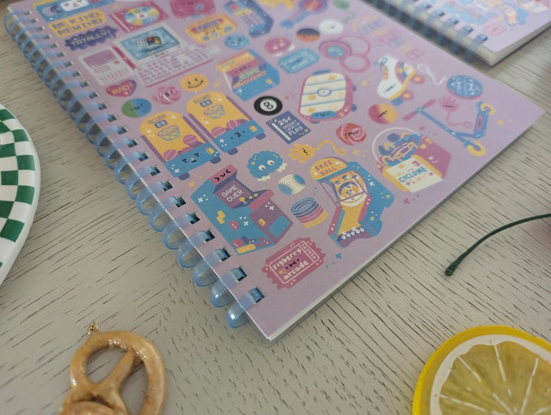 May include: A lilac spiral-bound notebook with a playful design of arcade game illustrations and smiley faces. The notebook has a clear plastic coil binding. The open pages are visible, showing the printed designs.