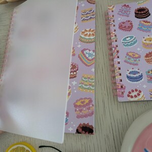 May include: A spiral-bound notebook with a lavender cover featuring colorful cake illustrations. The notebook is open, revealing a translucent cover sheet. A lemon slice and a cherry are also visible.