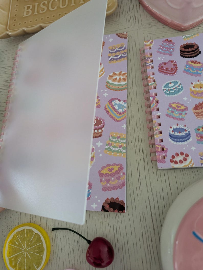 May include: A spiral-bound notebook with a lavender cover featuring colorful cake illustrations. The notebook is open, revealing a translucent cover sheet. A lemon slice and a cherry are also visible.