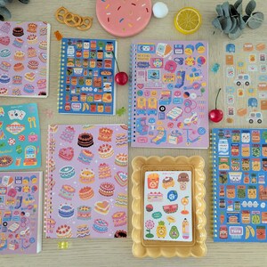 May include: A collection of vibrant notebooks and sticker sheets, adorned with designs of cakes, food items, and playful illustrations. The image also includes decorative elements such as cherries and lemon slices.