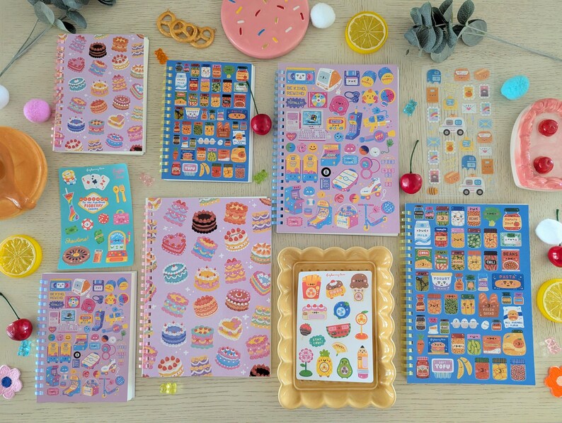 May include: A collection of vibrant notebooks and sticker sheets, adorned with designs of cakes, food items, and playful illustrations. The image also includes decorative elements such as cherries and lemon slices.