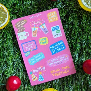 May include: A pink sticker sheet with various colorful bubble text designs, including "Cheers!", "Let's Hang Out!", and "Spend Time Together". The sheet is surrounded by artificial green grass, red berries, and lemon slices.