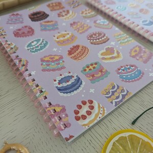 May include: A spiral-bound notebook with a lavender cover featuring colorful illustrations of various cakes. The cakes are decorated with frosting, berries, and other details. A second notebook with a similar design is visible.