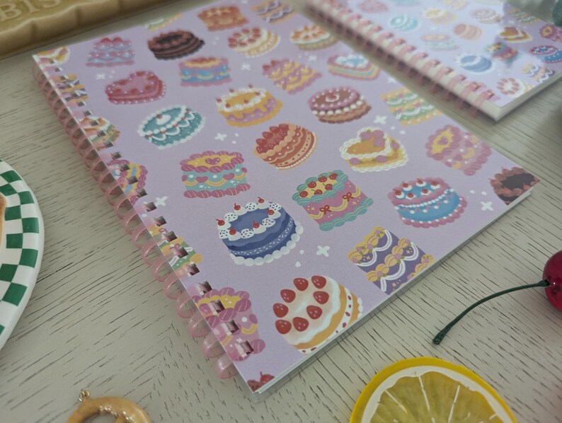 May include: A spiral-bound notebook with a lavender cover featuring colorful illustrations of various cakes. The cakes are decorated with frosting, berries, and other details. A second notebook with a similar design is visible.