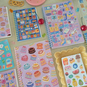 May include: A selection of vibrant spiral-bound notebooks and stickers. The notebooks display illustrations of food, cakes, and retro gadgets. A pink plate with sprinkles and a yellow picture frame are also present.