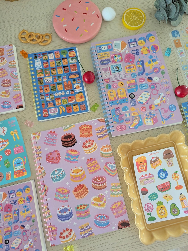 May include: A selection of vibrant spiral-bound notebooks and stickers. The notebooks display illustrations of food, cakes, and retro gadgets. A pink plate with sprinkles and a yellow picture frame are also present.