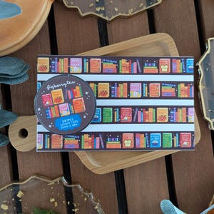 K&ouml;nnte beinhalten: Eine Rolle Washi-Tape und ein Streifen Washi-Tape mit einem farbenfrohen B&uuml;cherregal-Design. Die Tape-Rolle ist braun mit dem Text "figberrytea" und "BOOKS WASHI TAPE 15mm x 10m".