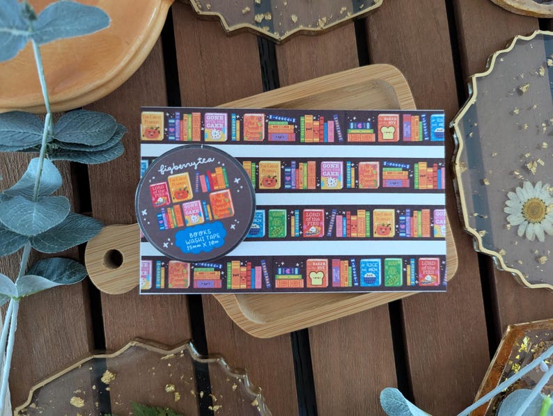 K&ouml;nnte beinhalten: Eine Rolle Washi-Tape und ein Streifen Washi-Tape mit einem farbenfrohen B&uuml;cherregal-Design. Die Tape-Rolle ist braun mit dem Text "figberrytea" und "BOOKS WASHI TAPE 15mm x 10m".