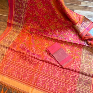 May include: A vibrant orange and pink textile featuring an intricate ikat pattern with geometric motifs and stylized animal figures. The fabric includes a wide decorative border with repeating patterns and a bottom edge finished with orange tassels.