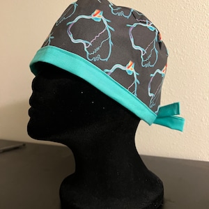 May include: A black surgical scrub cap with a teal trim and ties. The cap features a repeating pattern of colourful, abstract anatomical shapes. The trim and ties are a solid teal colour.