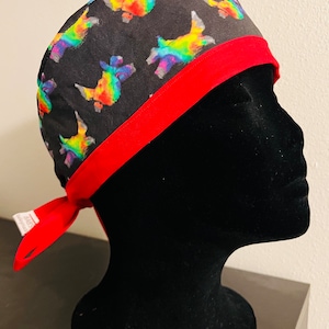 May include: A black surgical scrub cap with a repeating pattern of colorful, rainbow-colored dachshunds. The cap has a red band and ties in the back. The cap is displayed on a black mannequin head.