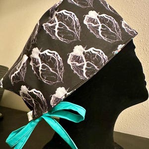 Könnte beinhalten: Eine schwarze OP-Haube mit einem sich wiederholenden Muster aus weißen anatomischen Herzillustrationen. Die Haube ist mit einer leuchtend türkisfarbenen Schleife gebunden. Die Haube ist für medizinisches Fachpersonal konzipiert.