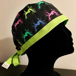 May include: Black surgical scrub cap with a neon green trim and ties. The cap is patterned with colorful, abstract tree-like designs. A small white tag with black text is attached to the tie. The cap is displayed on a black mannequin head.