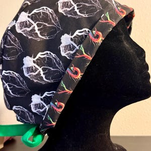 Könnte beinhalten: Schwarze OP-Haube mit weißem, venenartigem Muster. Der Rand der Haube weist ein farbenfrohes, abstraktes Design auf. Ein grünes Band sichert die Haube. Die Haube wird auf einem schwarzen Schaufensterkopf präsentiert.