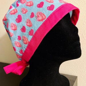 May include: A light blue scrub cap with a pattern of pink hearts and anatomical hearts. The cap has a bright pink trim and ties. The cap is designed for medical professionals.