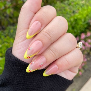 May include: Close-up of a hand with long, almond-shaped nails painted with a light pink polish. The tips of the nails are painted with a bright yellow accent and have small cherry designs.