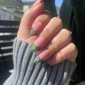 Könnte beinhalten: Nahaufnahme einer Hand mit langen, mandelförmigen Nägeln, die mit einem hellrosa Lack lackiert sind. Die Spitzen der Nägel sind mit einem grünen und schwarzen Blumendesign verziert.