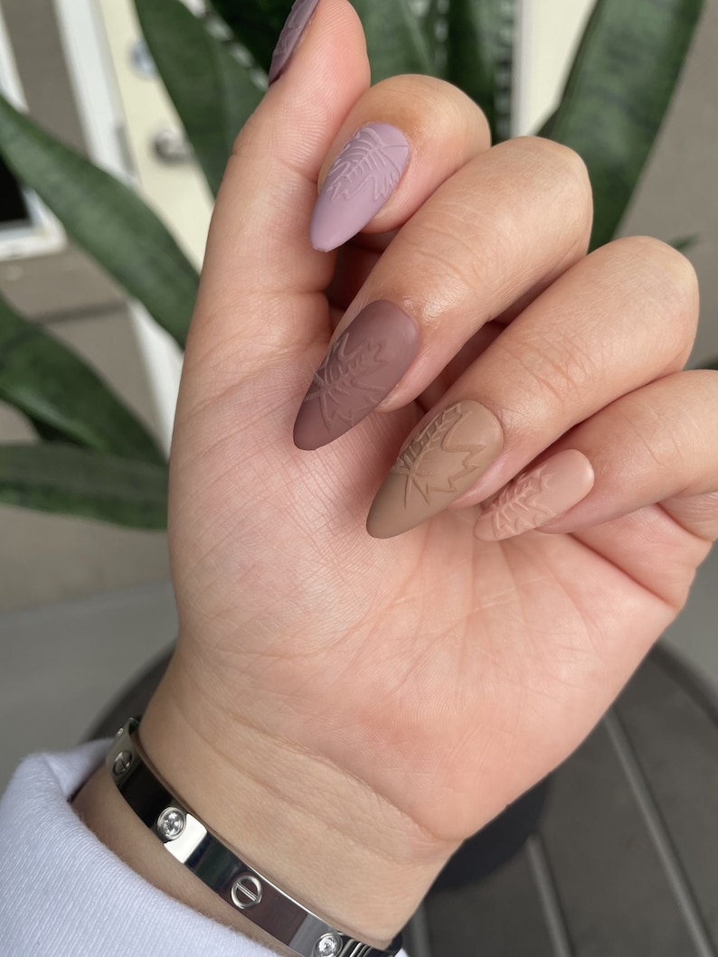 May include: Almond shaped nails with a matte finish. The nails are painted in shades of brown and purple with a leaf design embossed on each nail.