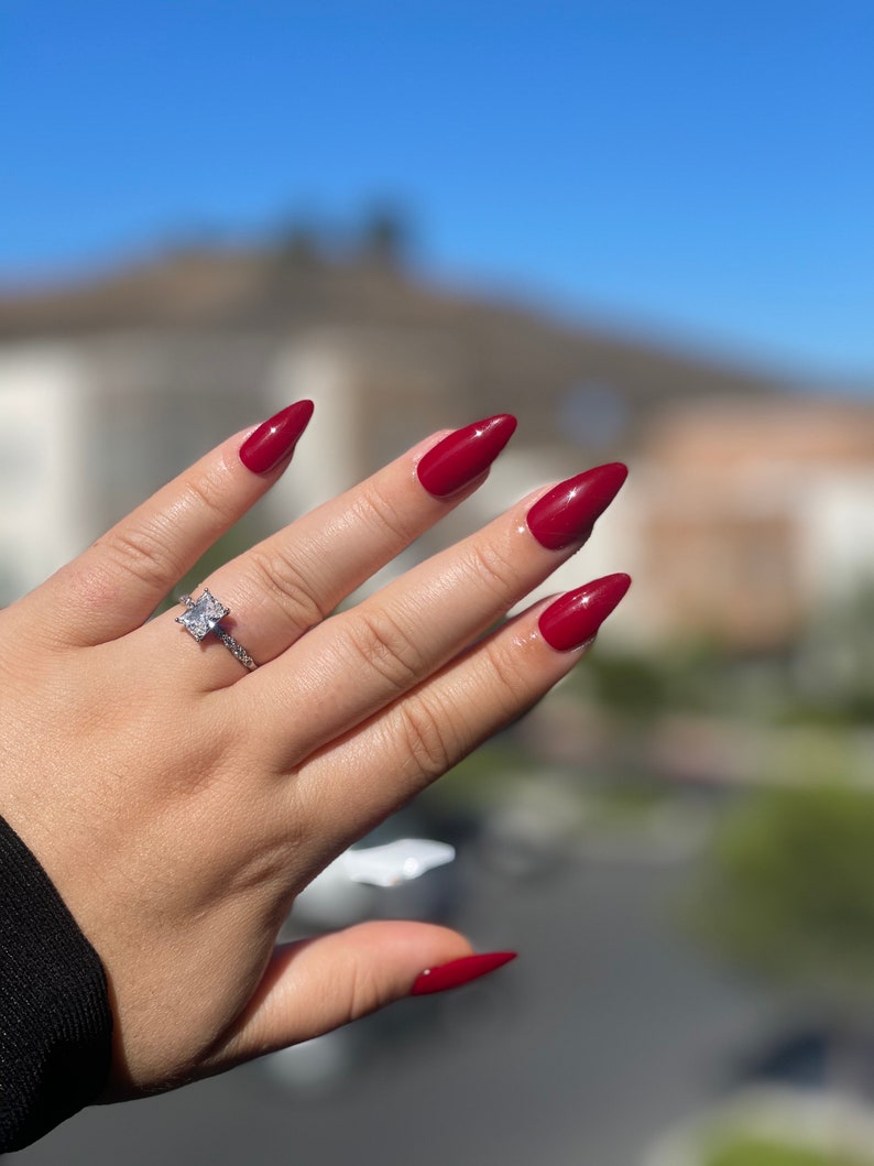 May include: Red stiletto nails with a clear square-shaped diamond ring on the ring finger.