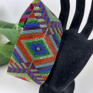Puede incluir: Una pulsera de cuentas coloridas con un patrón geométrico. La pulsera presenta una vibrante mezcla de colores que incluyen rojo, verde, azul y marrón. La pulsera se muestra en una exhibición negra en forma de mano.