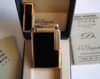 May include: A gold-plated S.T. Dupont lighter with a black rectangular panel, presented in its original navy blue box. The lighter features a rectangular design with gold detailing and a hinged lid. The box includes paperwork and the brand's logo.