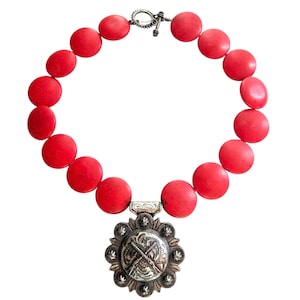 May include: A red beaded necklace with a silver pendant. The necklace features large, round red beads. The pendant is silver with a detailed design, including crossed rifles. The necklace has a toggle clasp.