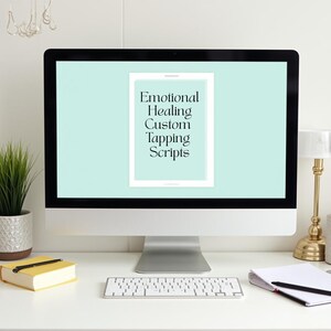 May include: A computer screen shows the text "Emotional Healing Custom Tapping Scripts" within a white frame on a light green backdrop. A yellow book, keyboard, and notepad are on the white desk.