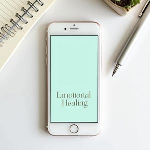 May include: A white smartphone with a gold-coloured frame displays the words "Emotional Healing" on a light green screen. A silver pen, a small potted plant, and a spiral notebook are also visible on a white surface.