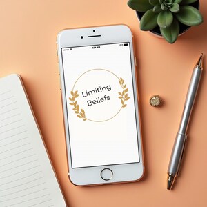 Może przedstawiać: Smartfon wyświetla tekst "Limiting Beliefs" w złotym wieńcu. Srebrny długopis, mały złoty pierścionek, sukulent i notatnik ułożone są na brzoskwiniowej powierzchni, tworząc scenę roboczą.