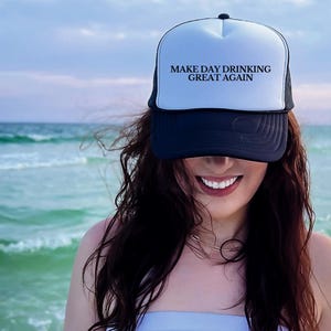 Könnte beinhalten: Eine schwarz-weiße Trucker-Kappe mit dem Text "Make Day Drinking Great Again" auf der Vorderseite. Die Kappe wird von einer Person mit langen braunen Haaren getragen, die vor einer Strandkulisse steht.