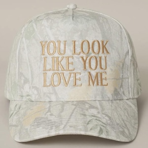 May include: A white baseball cap with a camouflage pattern and the text "YOU LOOK LIKE YOU LOVE ME" embroidered in gold.