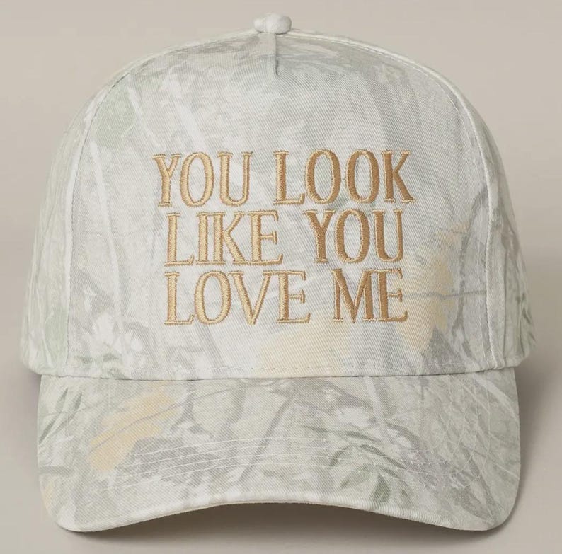 May include: A white baseball cap with a camouflage pattern and the text "YOU LOOK LIKE YOU LOVE ME" embroidered in gold.