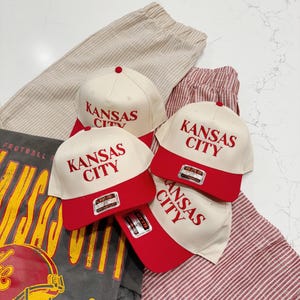 May include: Three white baseball caps with red brims and the text "Kansas City" in red lettering. The caps are on a striped fabric background.